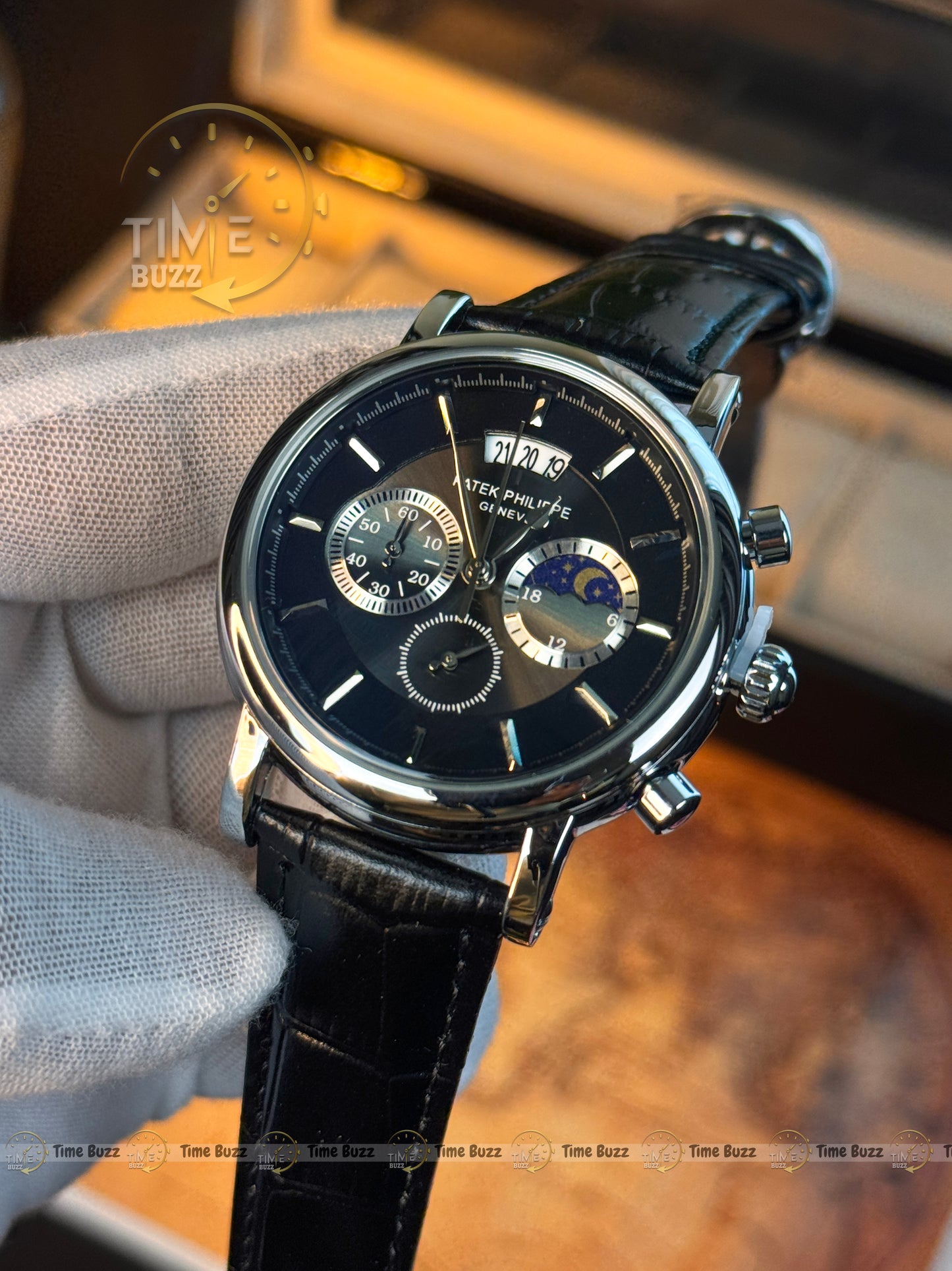 A Peter Millar GMT watch with a black dial, silver-tone case, and black leather strap, held in a white-gloved hand against a warm, blurred background.