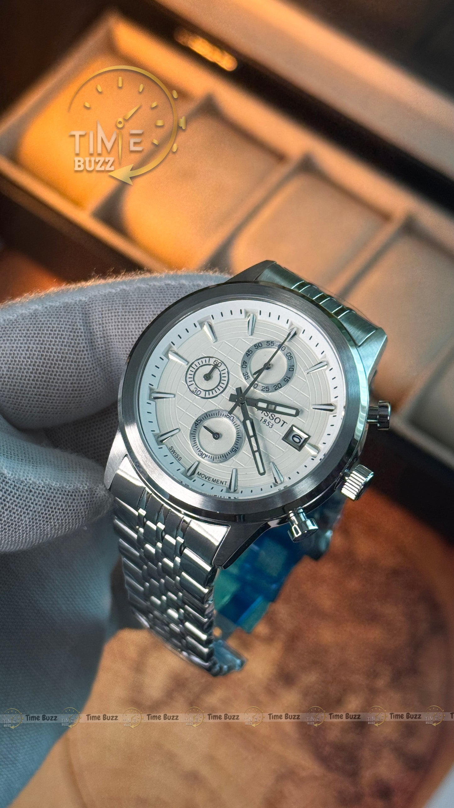 A silver stainless steel chronograph watch with a white dial and silver link bracelet, held by a hand wearing a gray glove. The watch has three sub-dials, a date window, and a blue crystal protective cover on the crown.