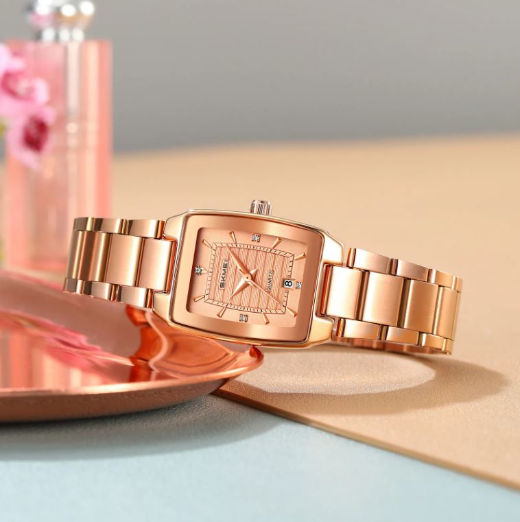 Rose gold watch on a light blue surface with a pink flower in the background