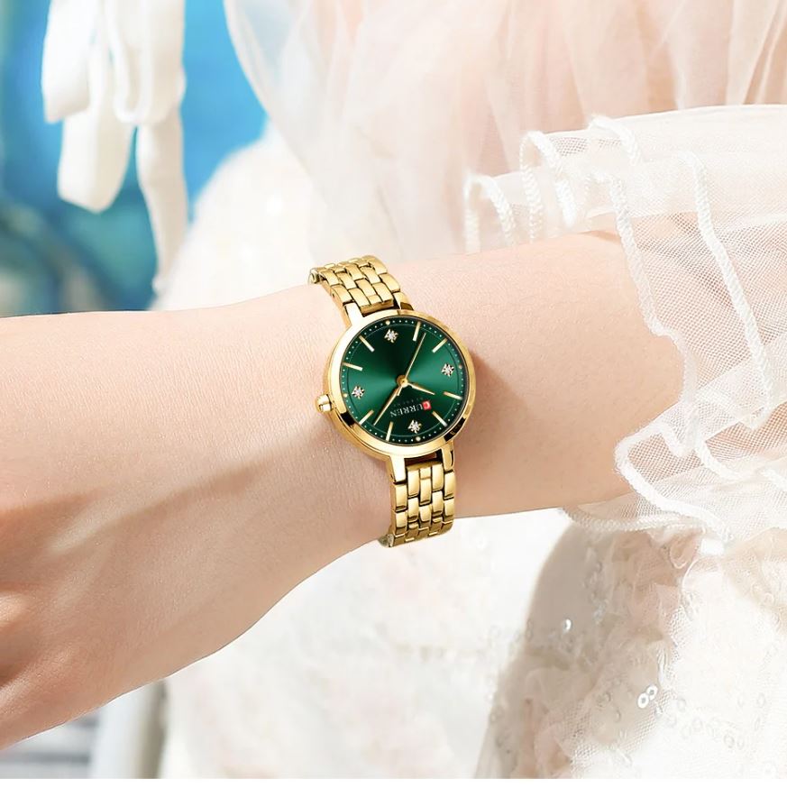 Gold watch with a green face on a wrist, wearing a white lace garment.