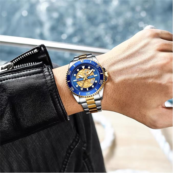 Wrist wearing a watch with a blue face and gold accents, with a blurred background.