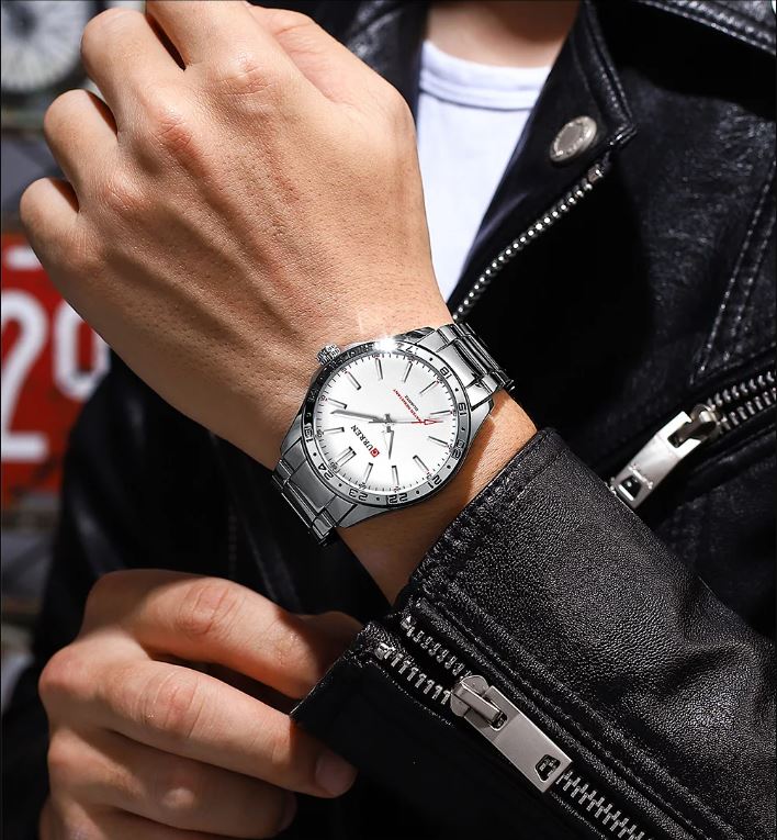 Hand wearing a silver watch with a white face, against a blurred background of a person in a black jacket.