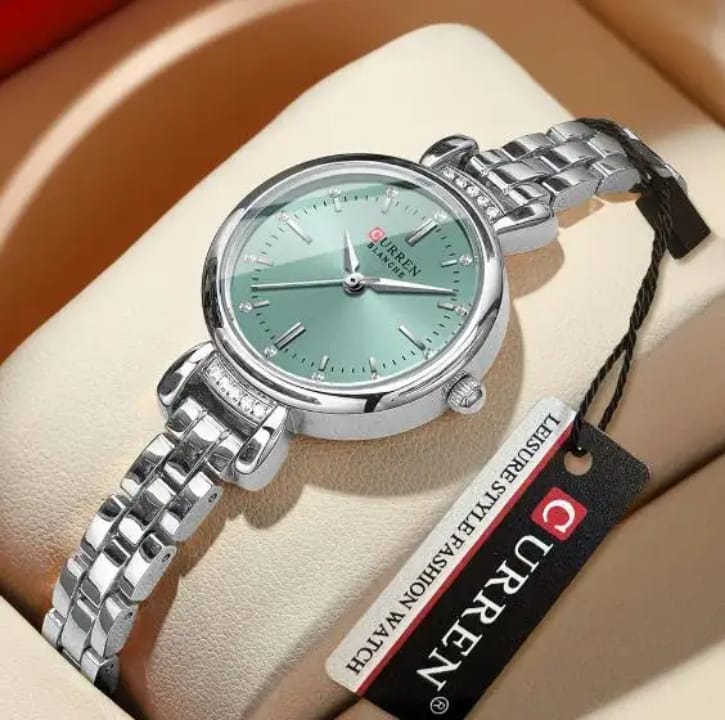 Silver watch with green face on a beige cushion, branded 'UREN'.